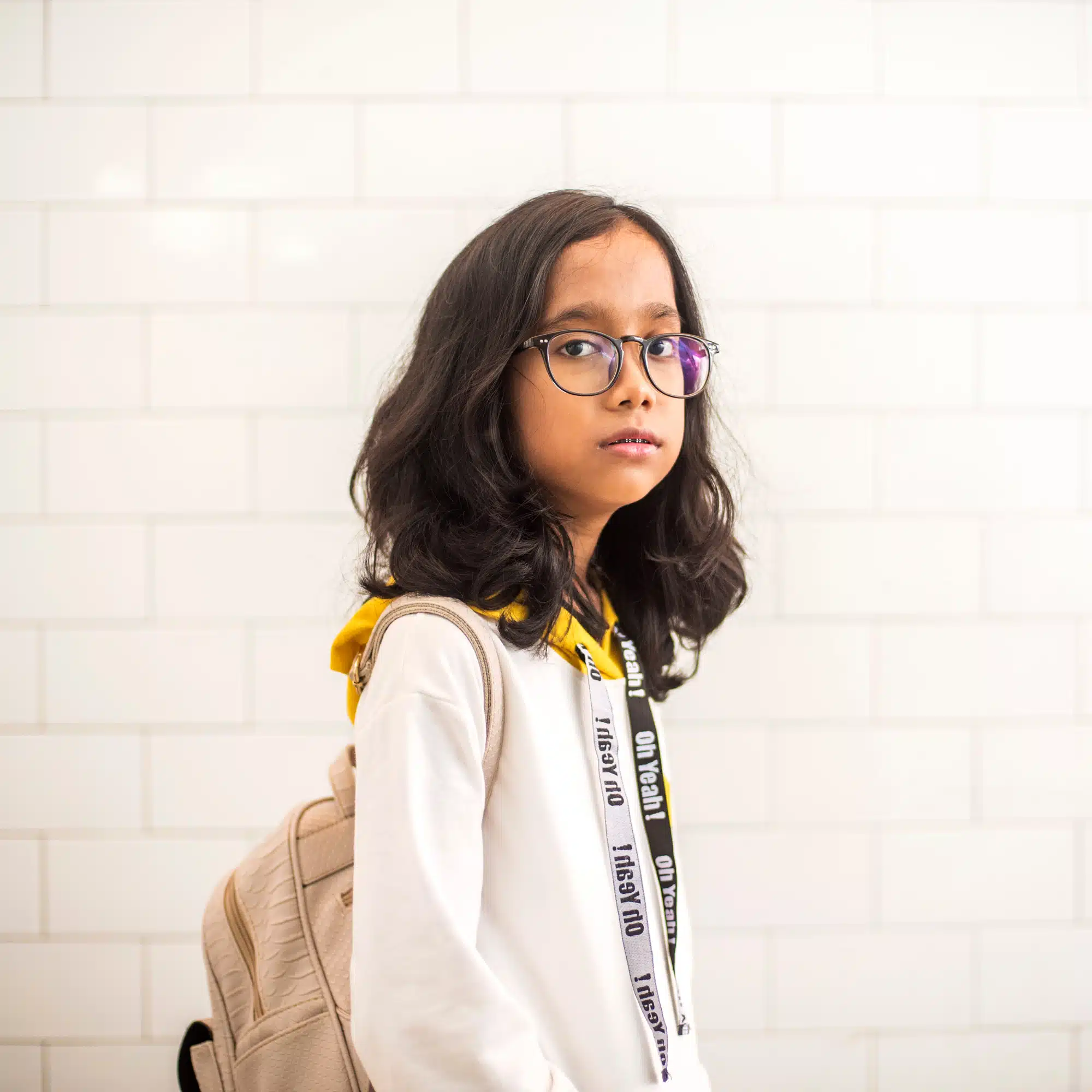 12 year old girl with glasses and backpack on standing in front of white brick wall